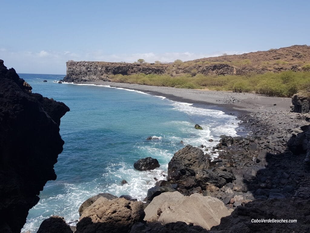 View of Aguas Belas Natural Cave on Santiago Island, Cape Verde, showing basalt cliffs, turquoise lagoon, and nearby beach under sunlight.