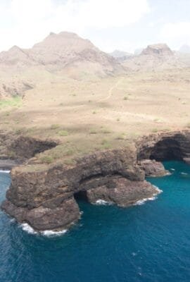 View of Aguas Belas Natural Cave on Santiago Island, Cape Verde, showing basalt cliffs, turquoise lagoon, and nearby beach under sunlight.