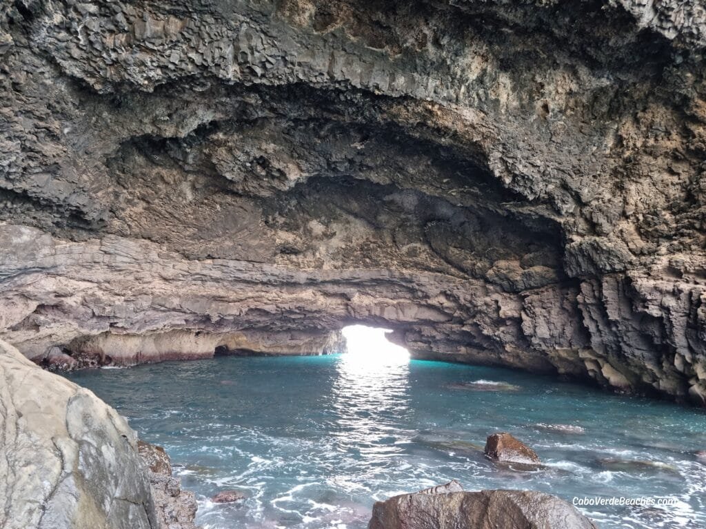 View of Aguas Belas Natural Cave on Santiago Island, Cape Verde, showing basalt cliffs, turquoise lagoon, and nearby beach under sunlight.