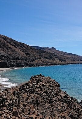 A panoramic view of Baixo Rocha Beach in São Nicolau, Cape Verde, showing a stretch of golden and black sand bordered by clear turquoise waters and rocky cliffs under a bright blue sky.