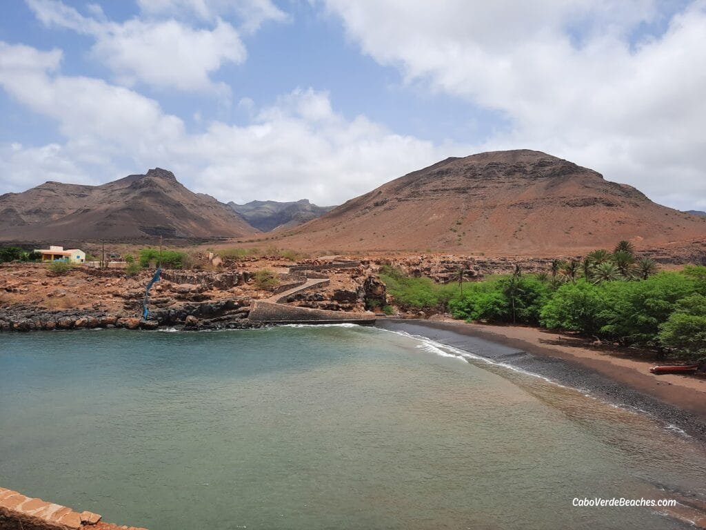 A quiet view of Baia Gombeza Beach in Cape Verde, with dark volcanic sand meeting the deep blue ocean, framed by rocky cliffs and a clear sky.