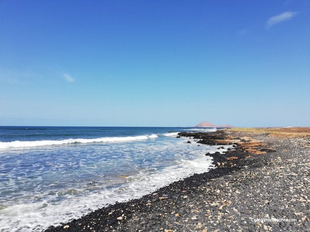 A dramatic view of Black Beach - Sal with black volcanic sand and rugged rock formations under a bright Cape Verde sky, waves gently rolling onto shore.