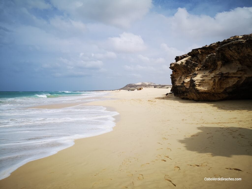 A panoramic view of the Beach of Varandinha in Cape Verde, showcasing golden sands, dramatic sea caves, and unique rock formations under a clear sky.