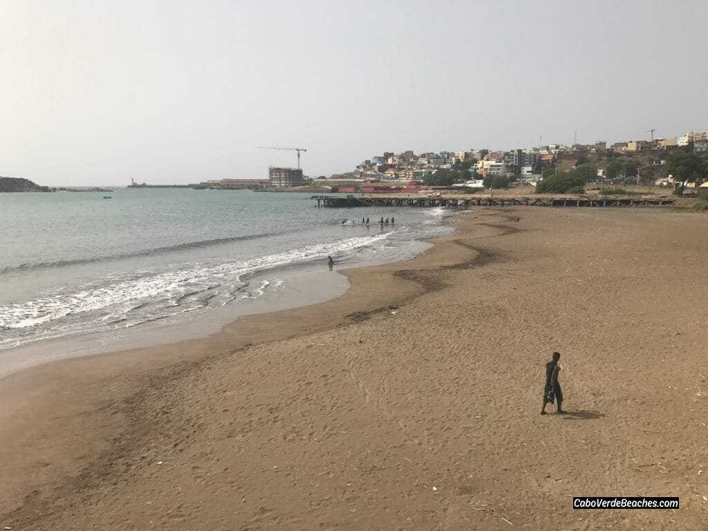 A panoramic view of Gamboa Beach in Praia, Santiago Island, showcasing its dark sandy shore, gentle waves, and the city's skyline in the background.