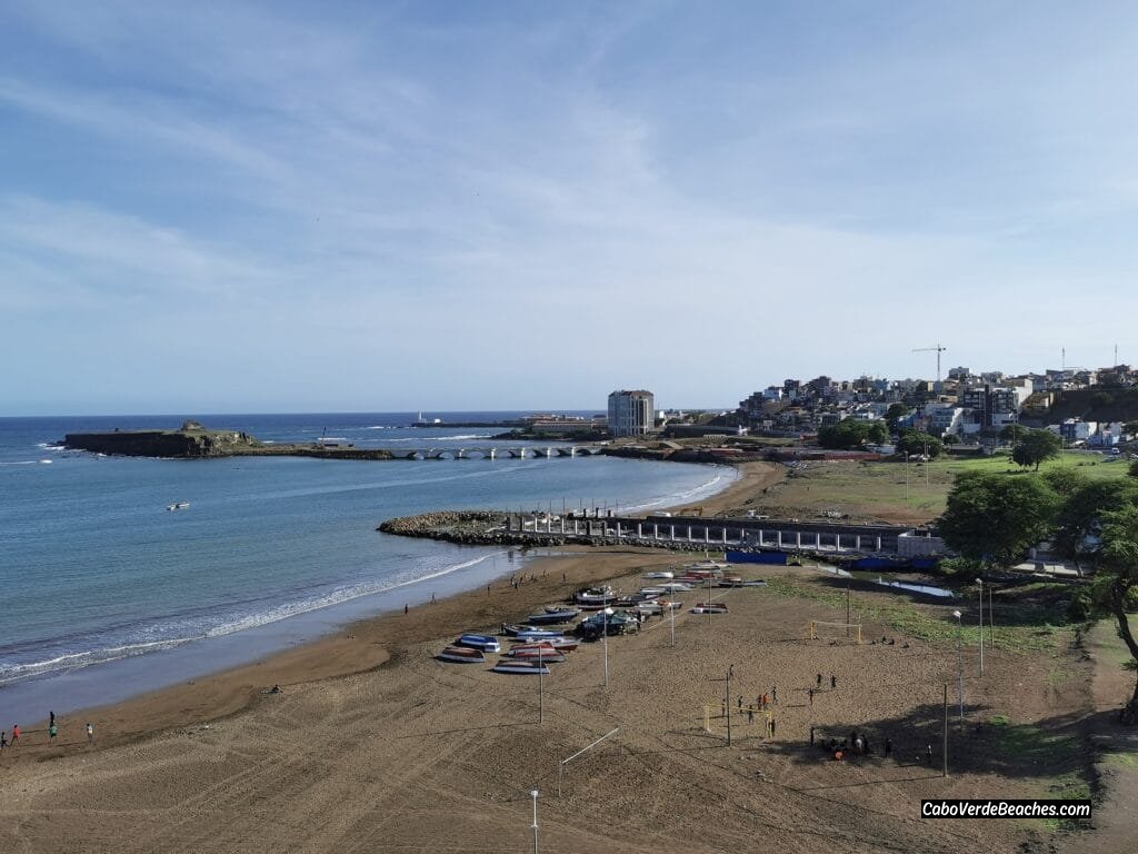 A panoramic view of Gamboa Beach in Praia, Santiago Island, showcasing its dark sandy shore, gentle waves, and the city's skyline in the background.