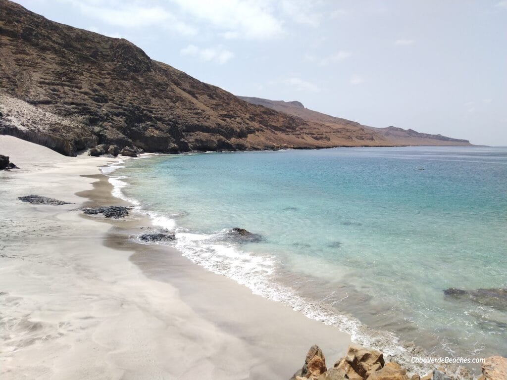 A panoramic view of Baixo Rocha Beach in São Nicolau, Cape Verde, showing a stretch of golden and black sand bordered by clear turquoise waters and rocky cliffs under a bright blue sky.
